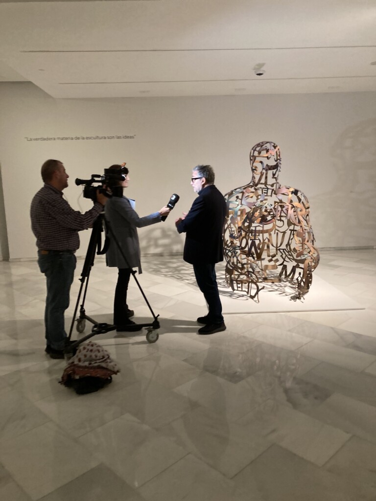 Image of journalists interviewing a man in front of a sculpture by Jaume Plensa