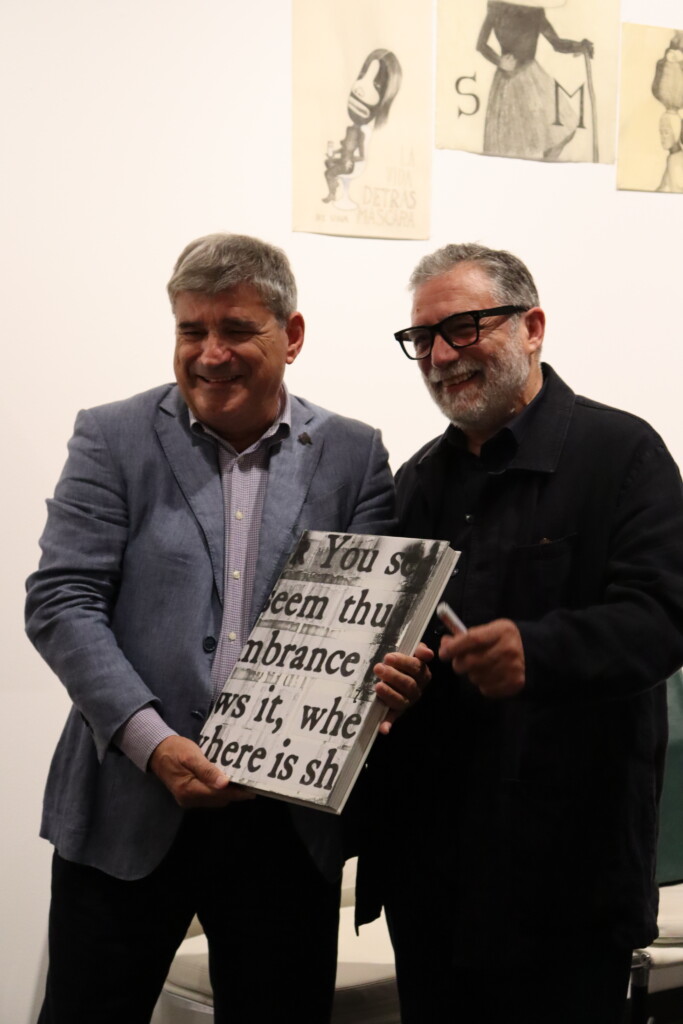 Fotografía de dos hombres sosteniendo un libro grande con letras negras sobre fondo blanco